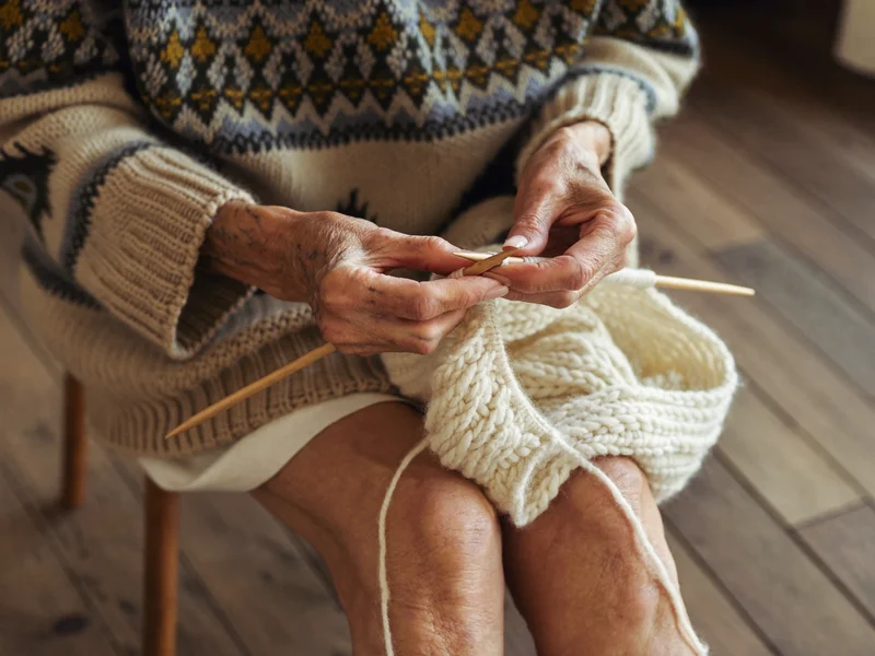 Mani anziane che lavorano a maglia con lana color crema e ferri di legno.